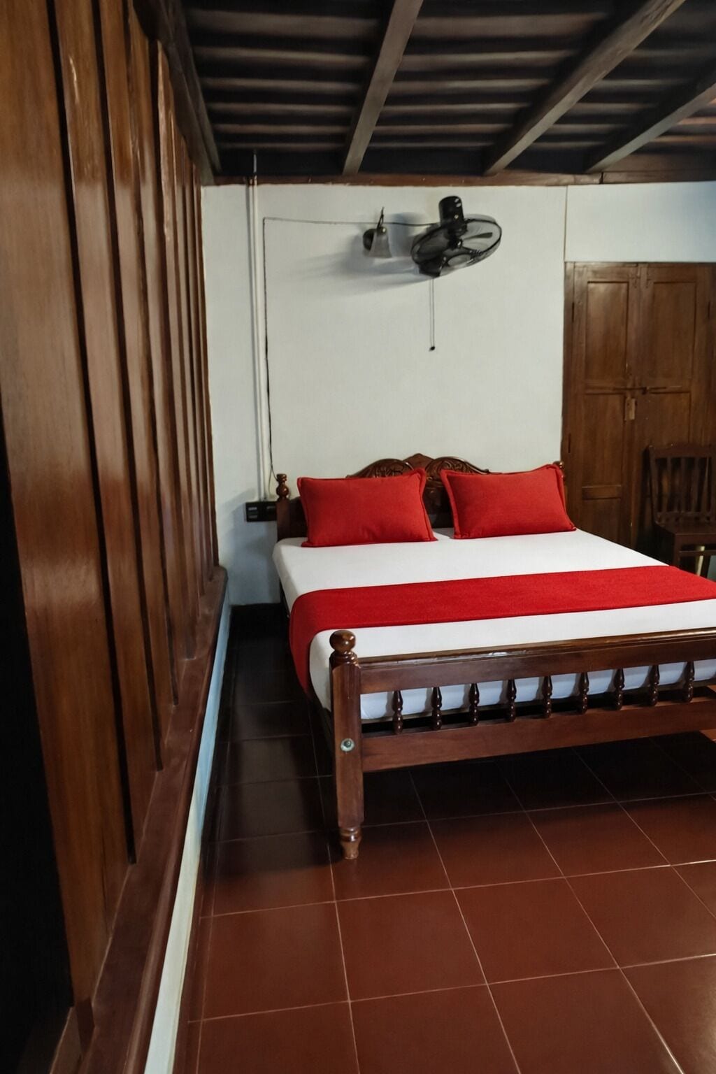 A bedroom with a wooden bed, red pillows, a white bedspread, a ceiling fan, and a wooden wardrobe.