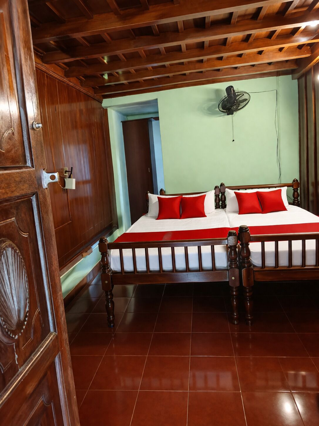 A wooden-framed bed with red and white bedding, a fan on the wall, and a wooden door with a handle.