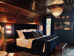 Una camera da letto accogliente con soffitto in legno, un letto grande con cuscini a motivi, una panca, una TV a parete e un lampadario.