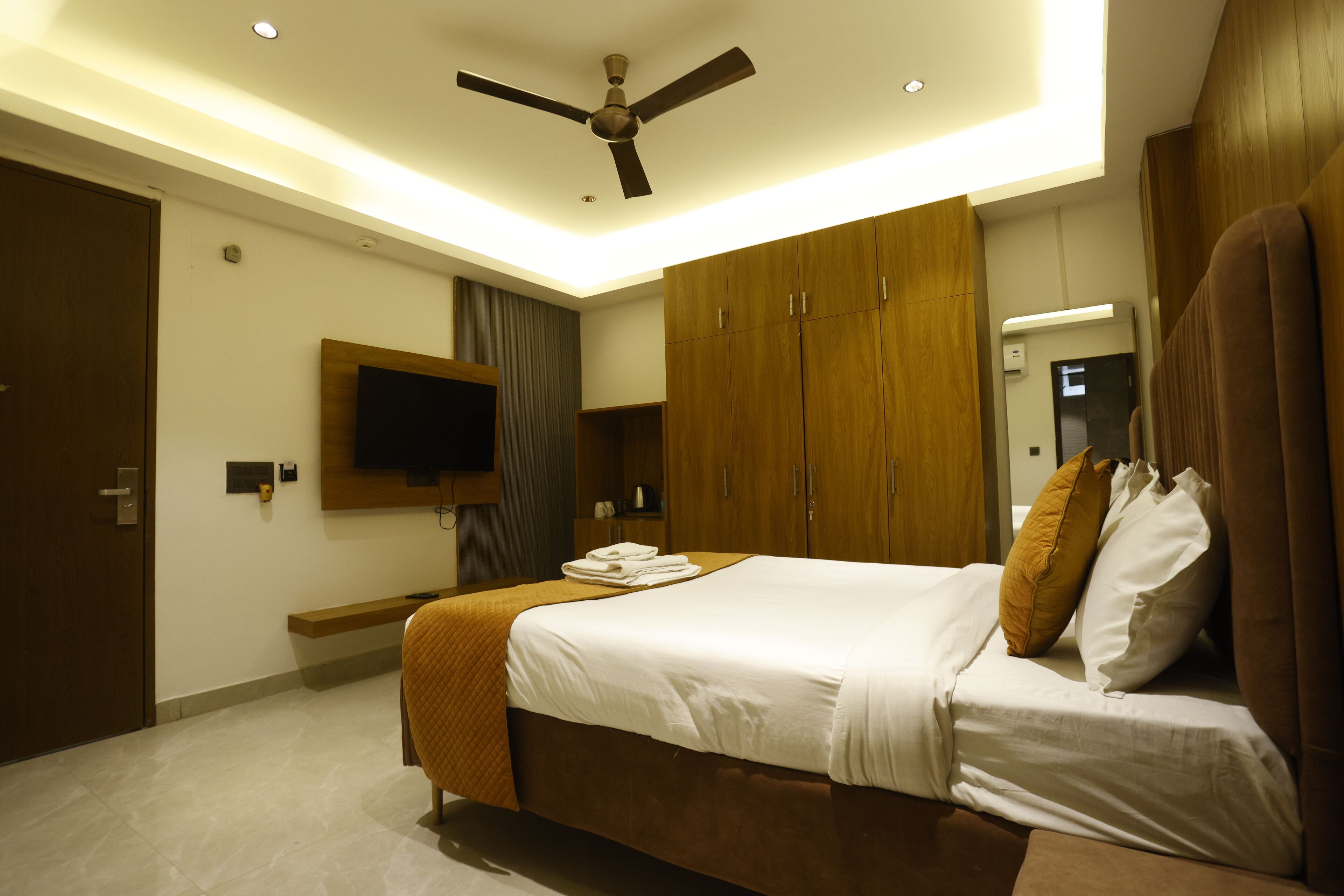 A modern hotel room with a bed, wooden wardrobe, a flat-screen TV, and a ceiling fan.