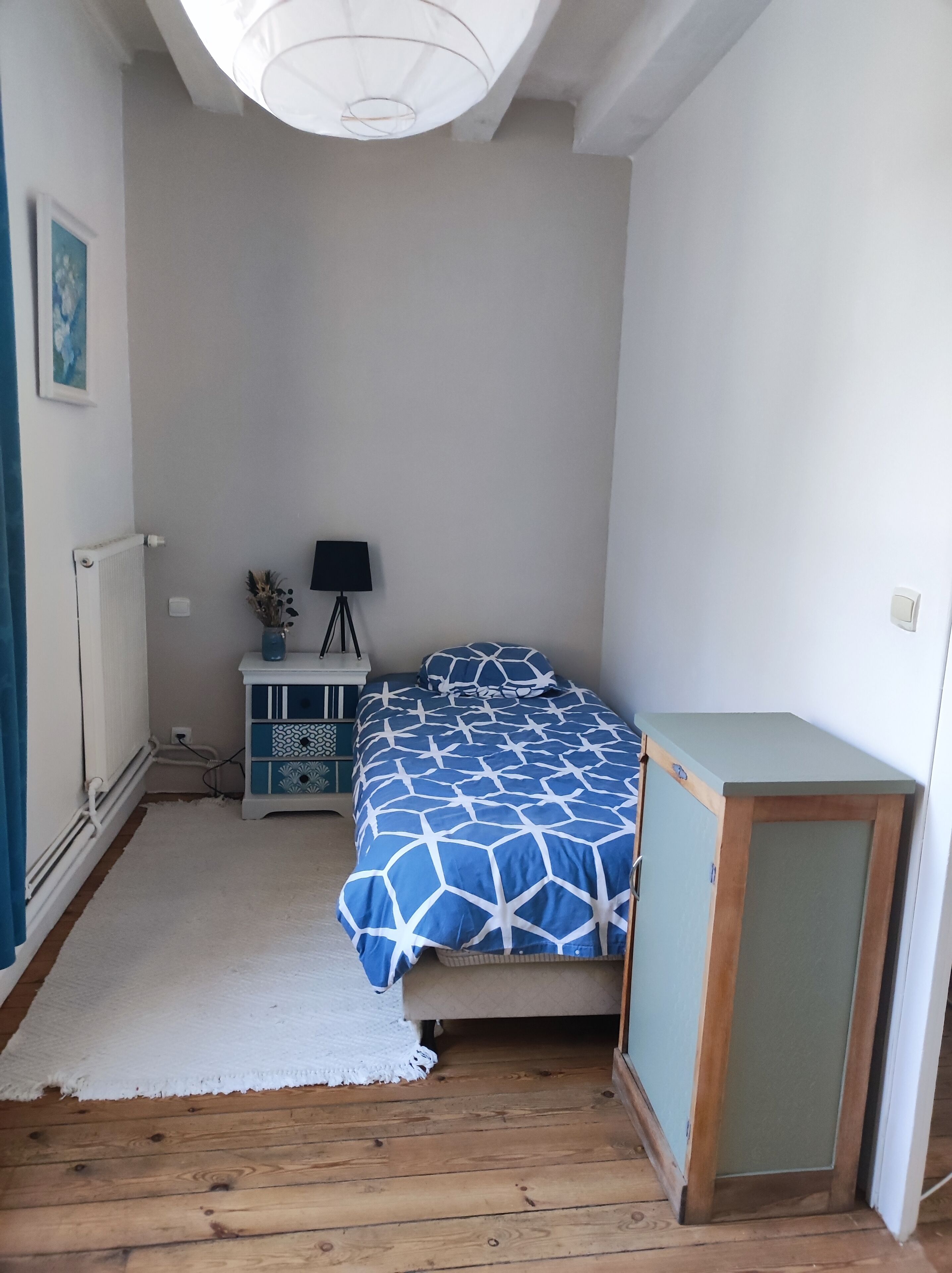 Et soveværelse med en seng, et natbord, en lampe, en radiator og et lille skab.