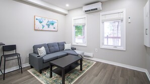 A modern living room with a grey sectional sofa, a dark coffee table, and a patterned rug. There are two windows with blinds, an air conditioning unit, and a world map artwork on the wall.