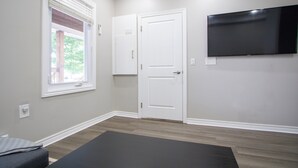 A room with a large flat-screen TV mounted on the wall, a window with blinds, a door, and a dark-colored exercise mat on the floor.