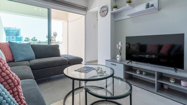 Ein modernes Wohnzimmer mit grauem Sofa, einem Glastisch, einem Flachbildfernseher und einem Balkon mit Blick auf Gebäude und Palmen.