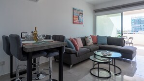 Ein modernes Wohnzimmer mit Essbereich, einem grauen Sofa, einem Glastisch und einem großen Fenster mit Blick auf die Stadt.