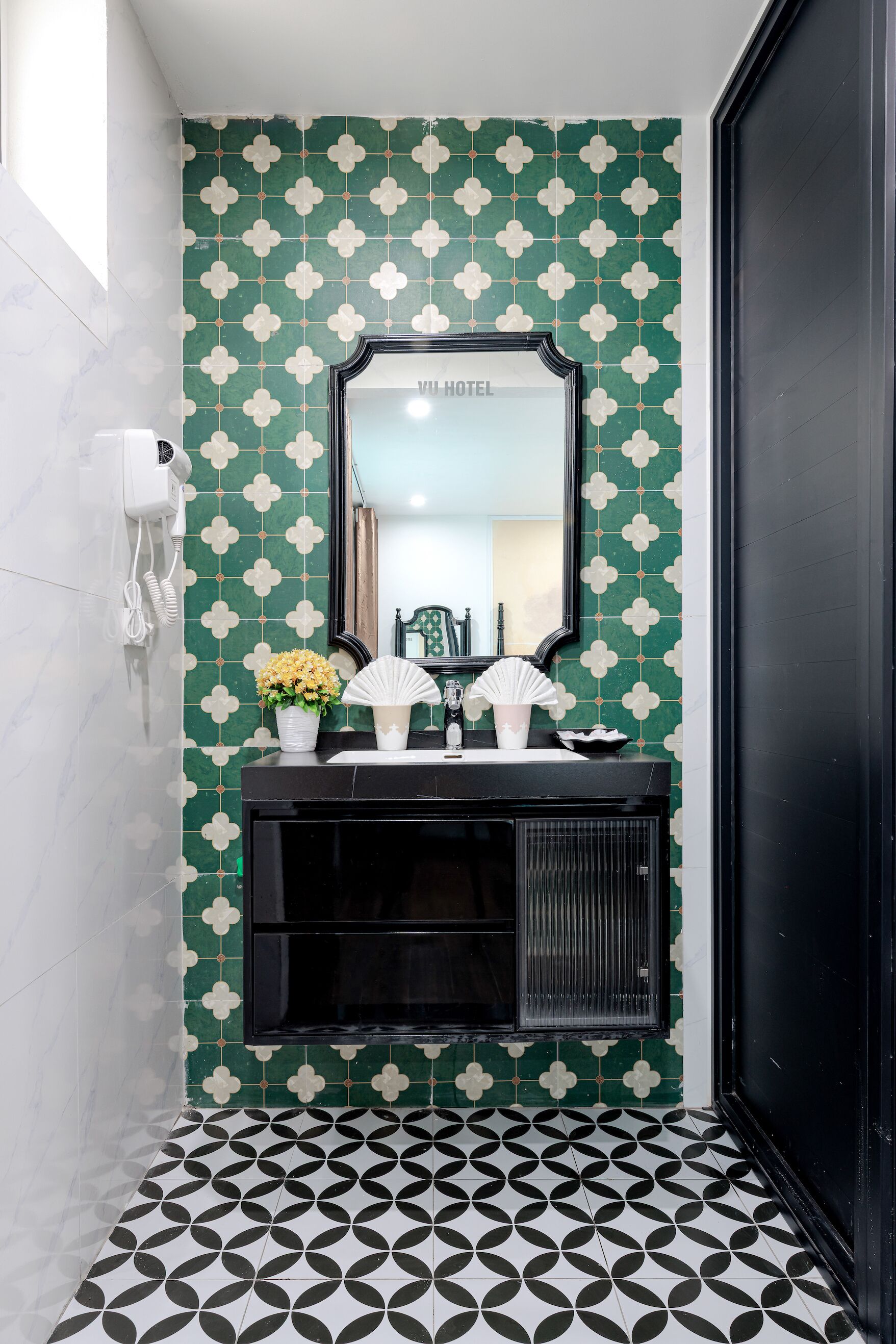Un baño con pared de azulejos blancos y verdes con motivos florales, un tocador negro con espejo, y piso con estampado blanco y negro.