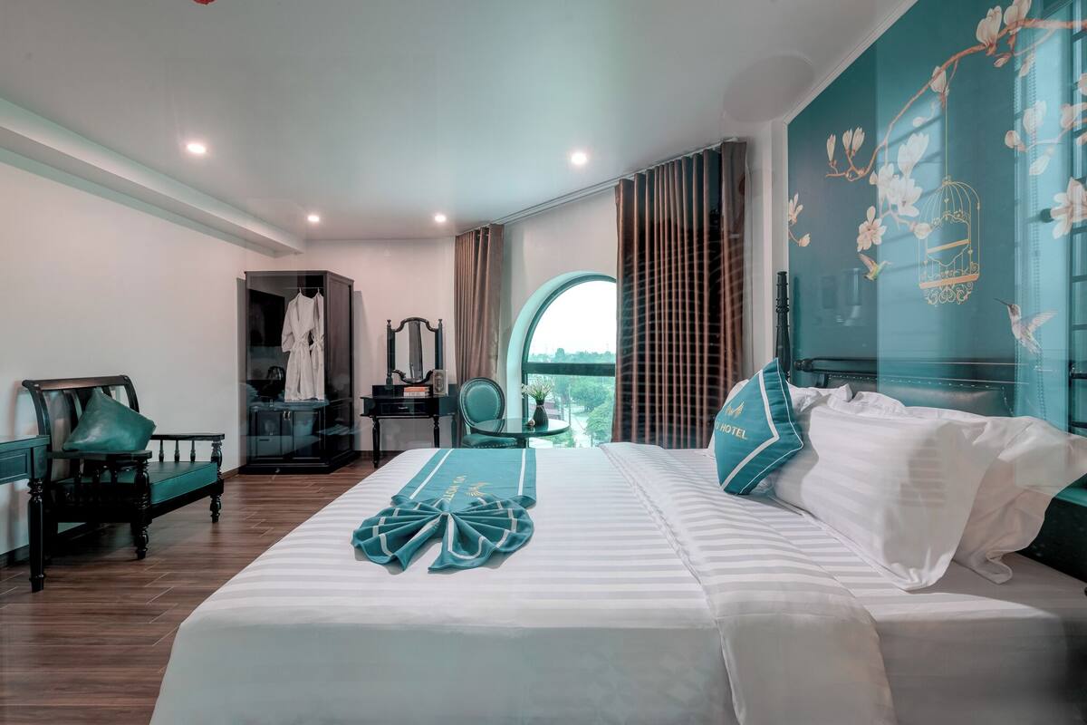 A hotel room with a large bed, a wardrobe, a vanity with a mirror, and a view of the outdoors through a window.