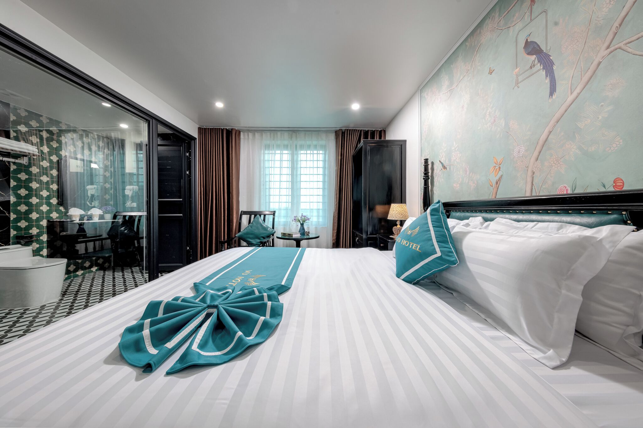 Quarto de hotel com uma cama grande, uma escrivaninha e uma cadeira. Há uma janela com cortinas, uma parede com um mural e um abajur na mesa de cabeceira.