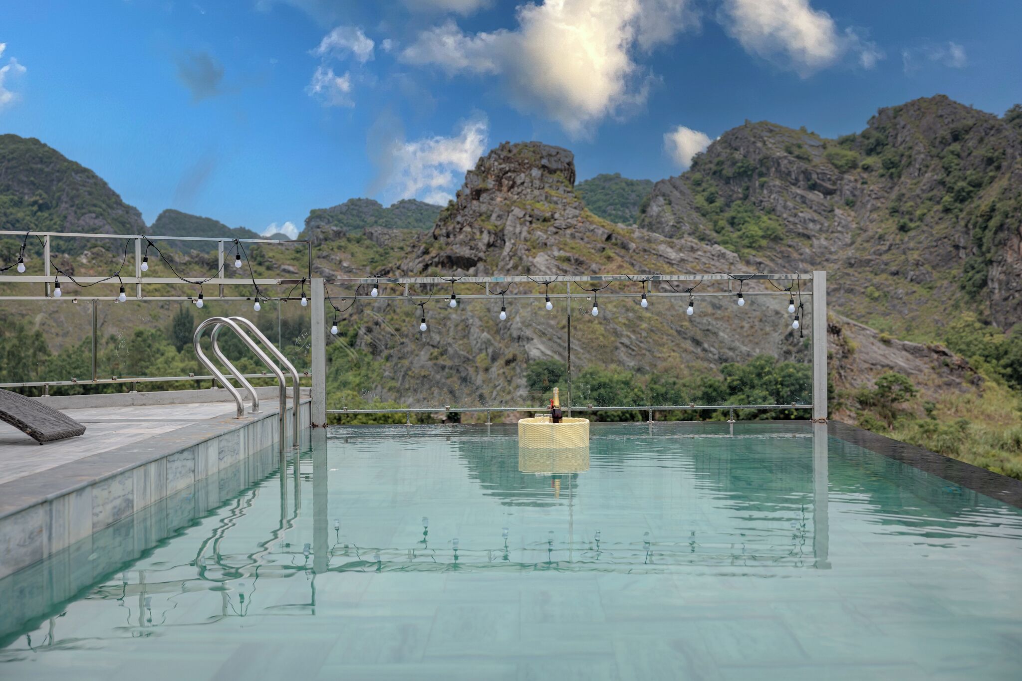 Una piscina con barandilla metálica, borde de piedra y montañas al fondo.