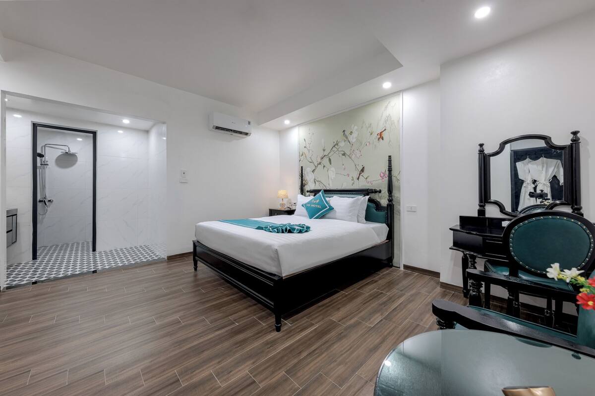 A modern hotel room with a large bed, a shower, a vanity with a mirror, and a wooden floor.