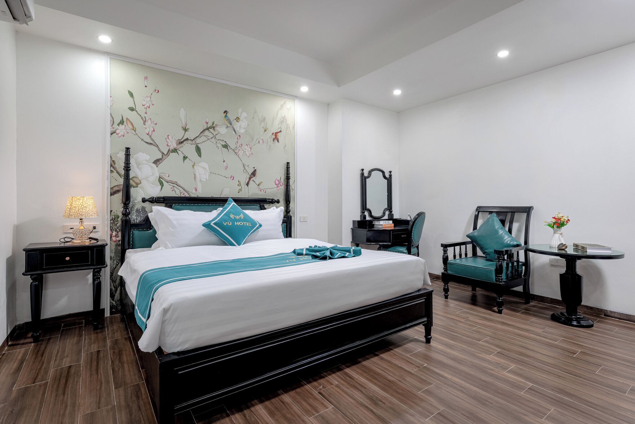 A hotel room with a bed, a desk, a chair, a small table, and a wall with a floral mural.