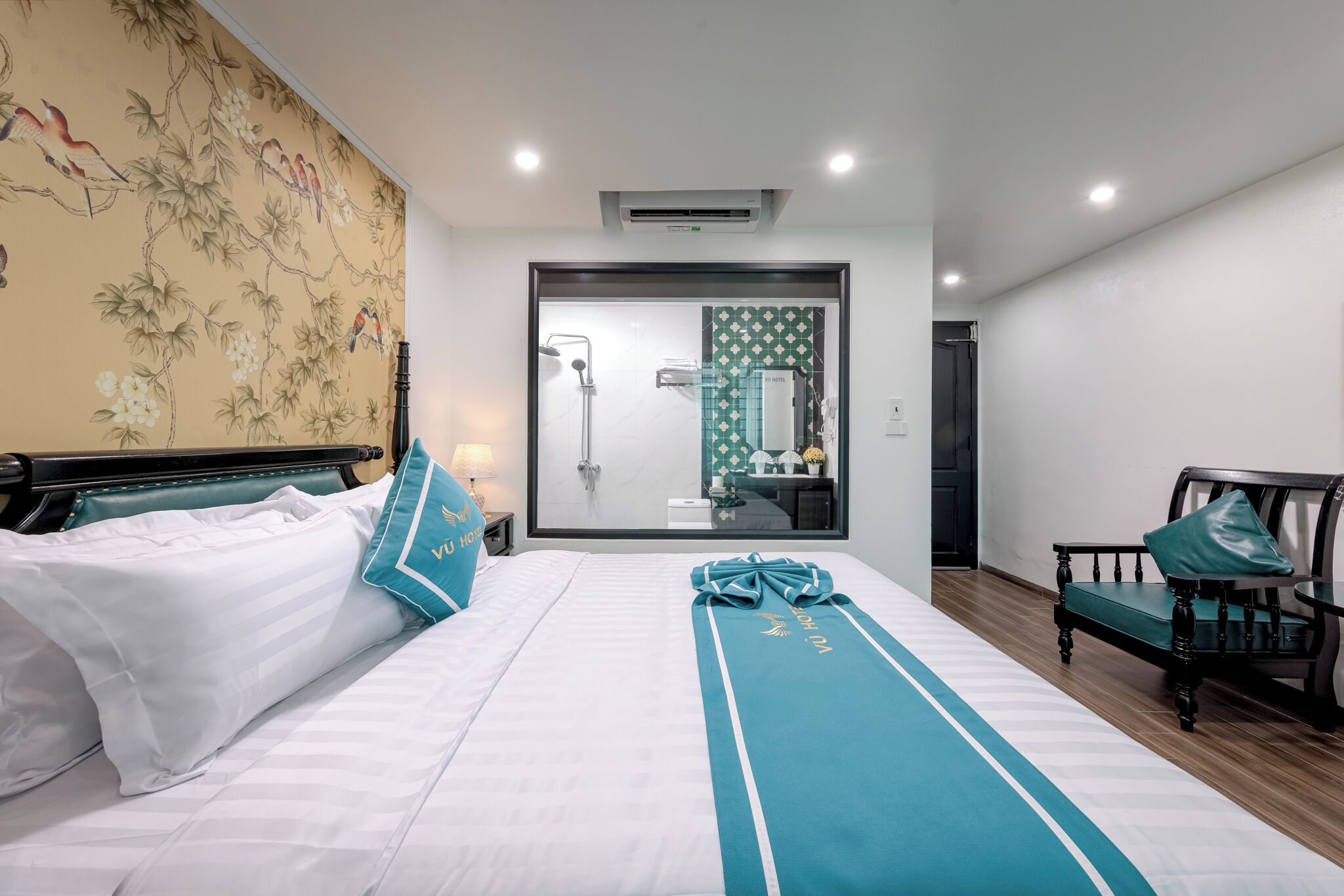 Une chambre d’hôtel avec un lit, un banc, un miroir et une salle de bain visible à travers une porte en verre.