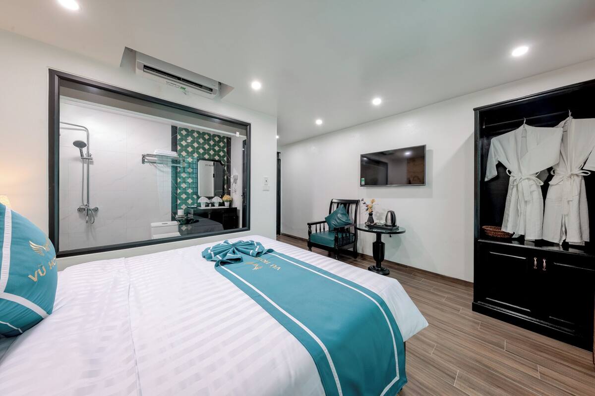 A hotel room with a bed, a shower, a vanity with a mirror, a TV mounted on the wall, and a wardrobe with clothes.