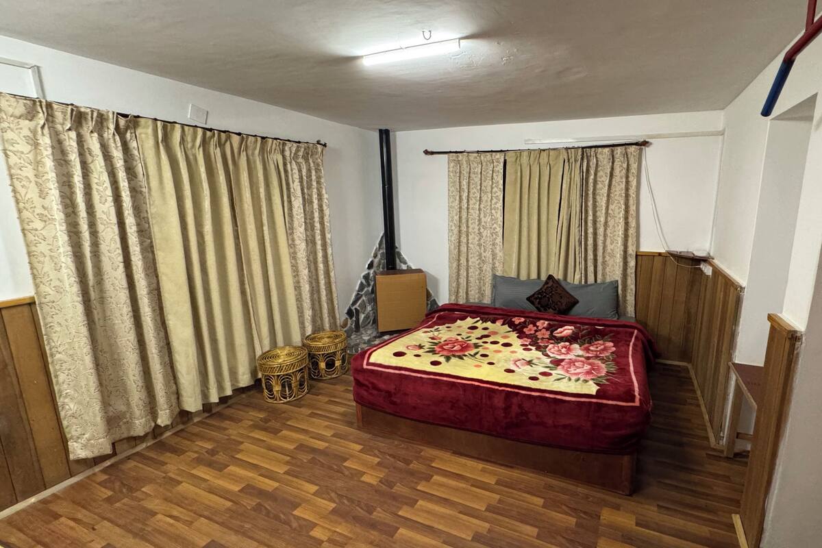 Quarto com piso de madeira, cama com colcha florida, duas almofadas, cabeceira de madeira e cortinas.