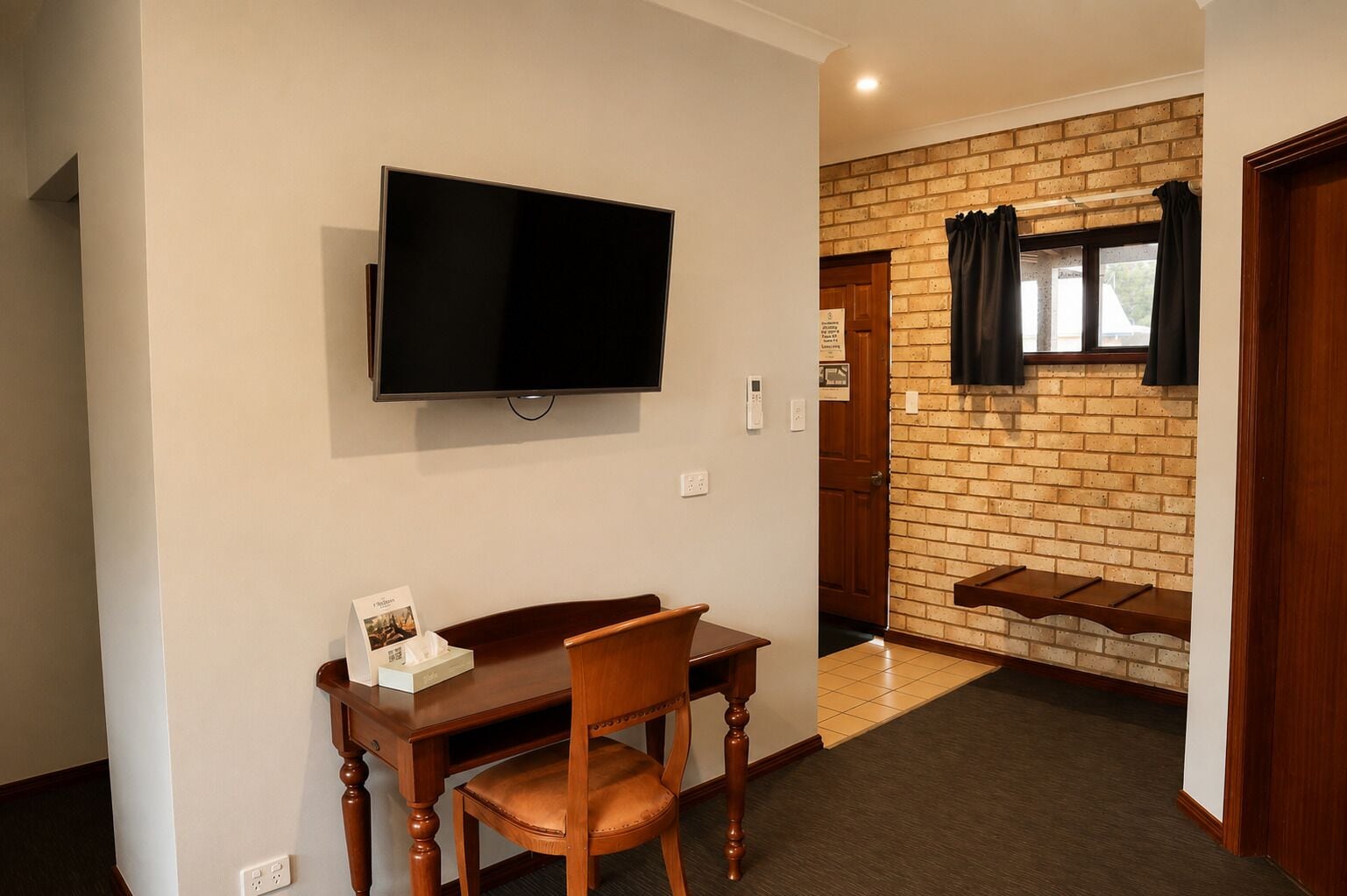 Ein Zimmer mit an der Wand befestigtem Fernseher, einem Holzespeisetisch mit einem Stuhl und einer Ziegelwand mit Fenster und Vorhängen.