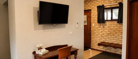 Ein Zimmer mit an der Wand befestigtem Fernseher, einem Holzespeisetisch mit einem Stuhl und einer Ziegelwand mit Fenster und Vorhängen.