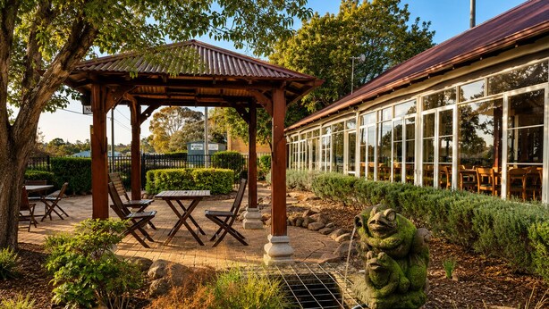 Un belvédère avec une table et des chaises, entouré de verdure et agrémenté d’une statue de grenouille en pierre.