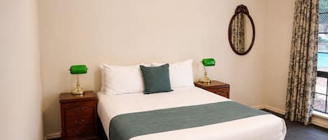Una cama bien hecha con un plaid verde, dos mesitas de noche con lámparas, un espejo y una ventana con cortinas.
