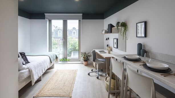 A modern, compact bedroom with a bed, a desk, and a view of the city through large windows. - Modern Studios with Kitchen & Ensuite (Edinburgh)