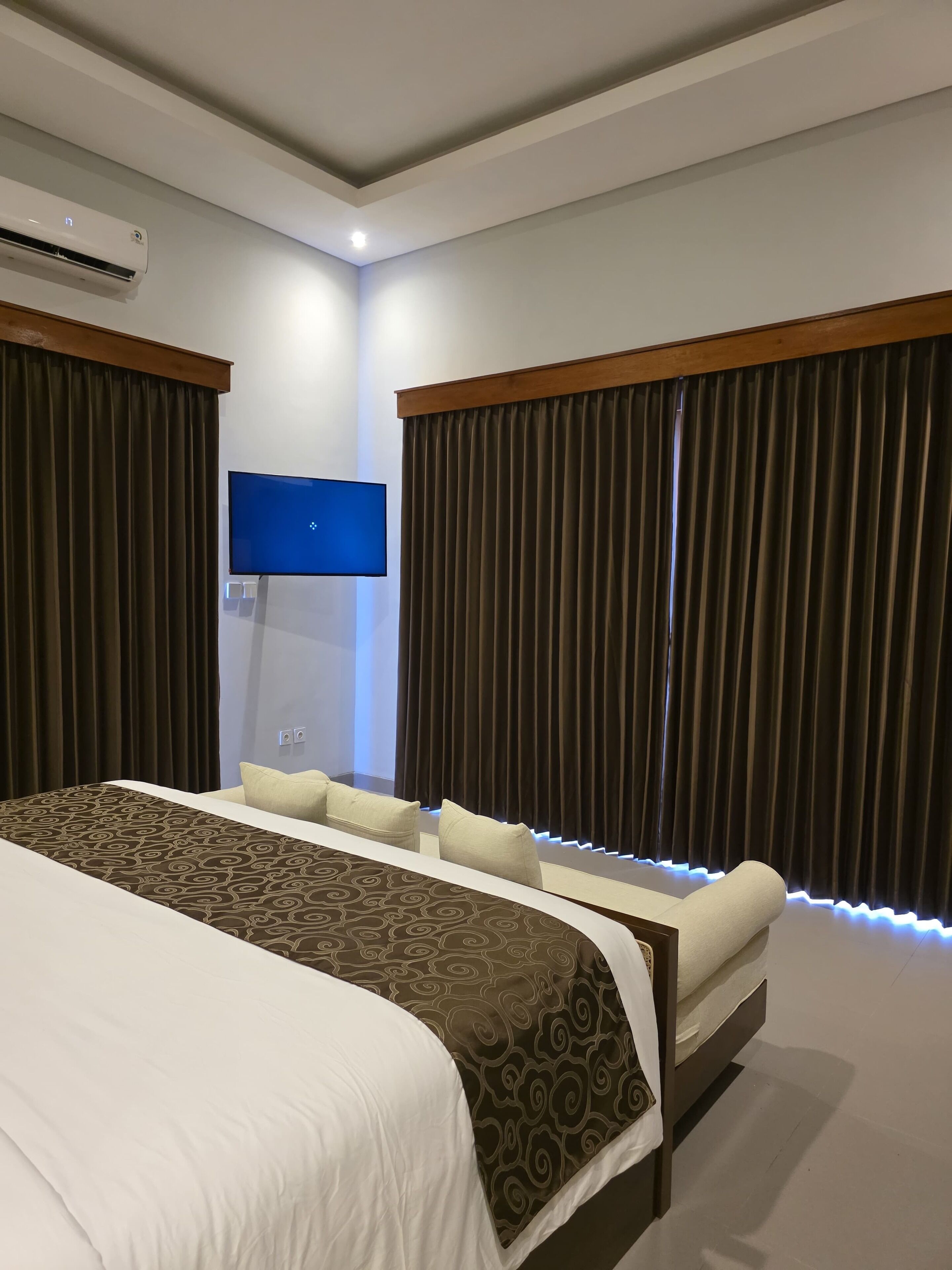 A modern hotel room with a large bed, a flat-screen TV mounted on the wall, and dark curtains on the windows.