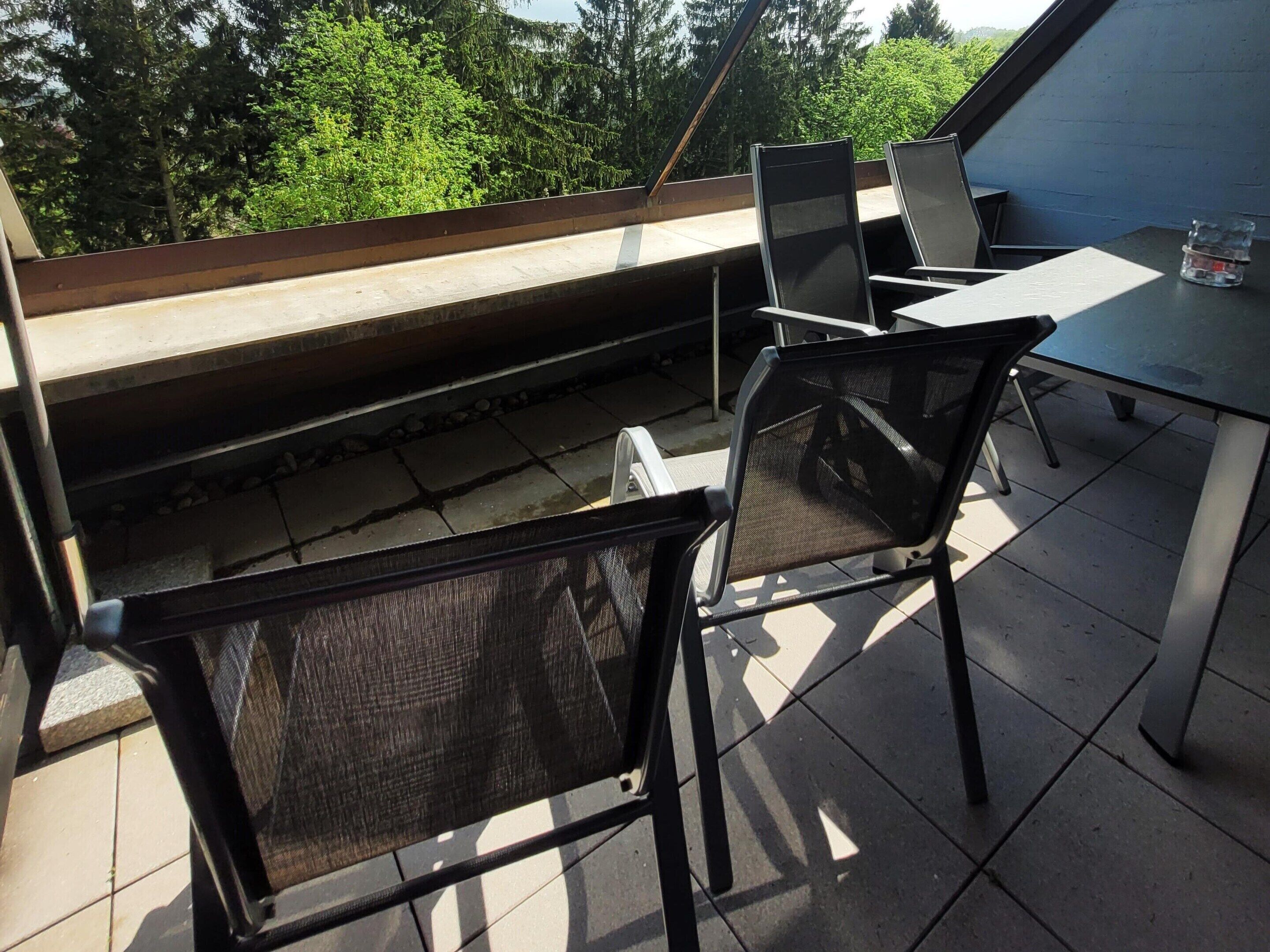 An outdoor patio area with a table and chairs, a view of trees, and a blue building in the background.