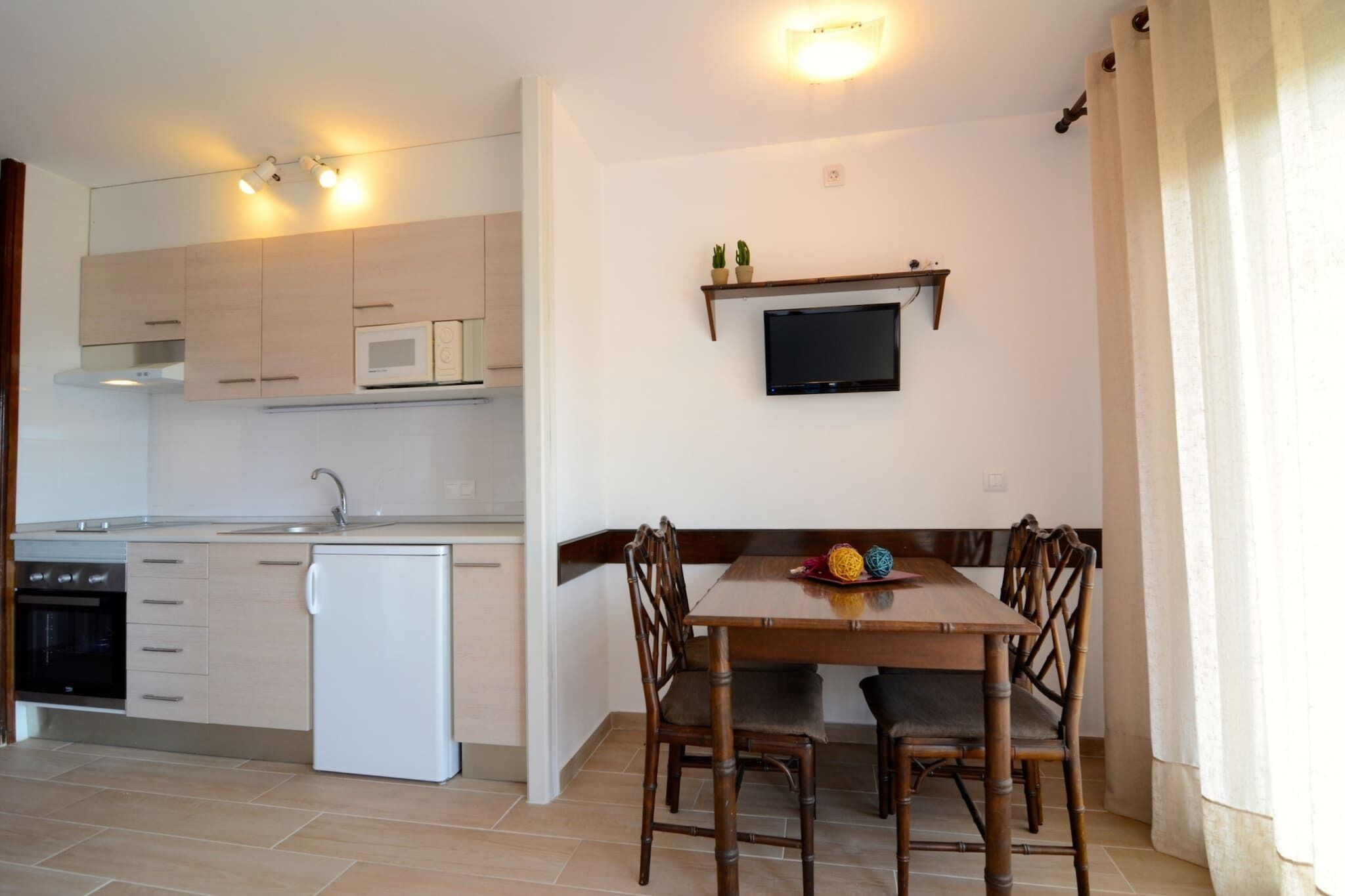 A compact kitchen with a microwave, oven, and dishwasher. There is a dining table with chairs and a flat-screen TV mounted on the wall.