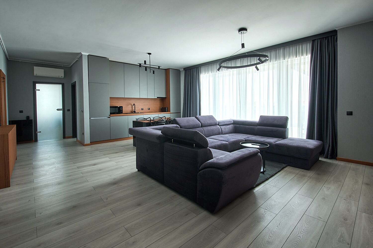 A modern living room with a sectional sofa, a dining area with a kitchen in the background, and a large window with curtains.