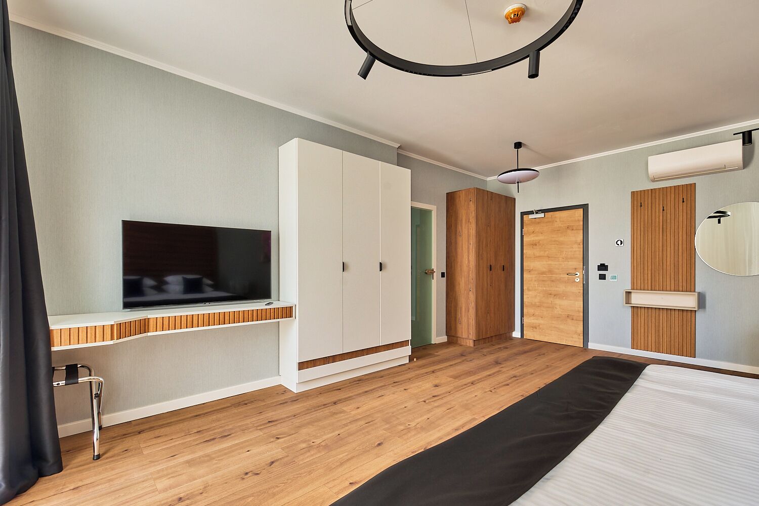 A modern hotel room with a large bed, a TV on a built-in shelf, a wardrobe, and a wardrobe mirror.