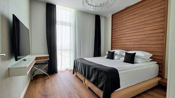 A modern bedroom with a wooden headboard, a flat-screen TV, a bedside table, and a large window with sheer curtains.