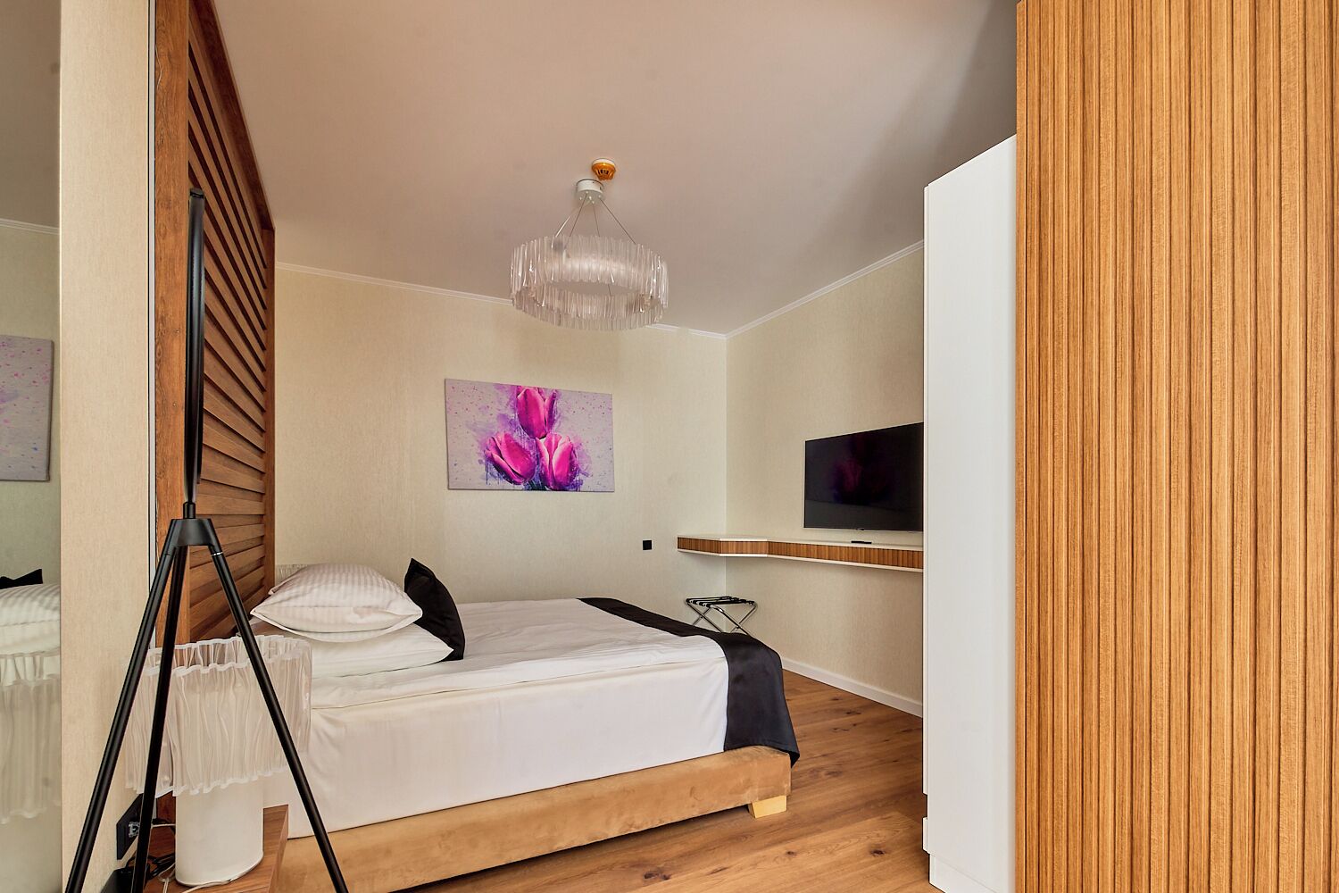 A bedroom with a bed, a television, a painting, and a chandelier.