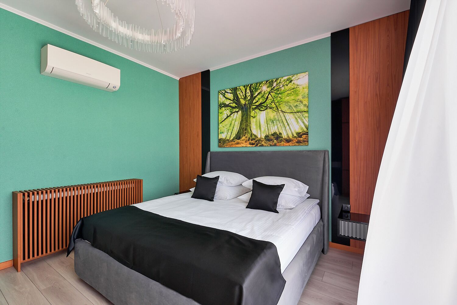 A modern hotel room with a bed, a radiator, a wall-mounted air conditioner, and a large nature-themed painting.