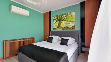A modern hotel room with a bed, a radiator, a wall-mounted air conditioner, and a large nature-themed painting.