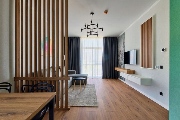 A modern living room with a wooden dining table, a sofa, a TV mounted on the wall, and a large window with curtains. - Bach Apartments 2 (Sibiu)