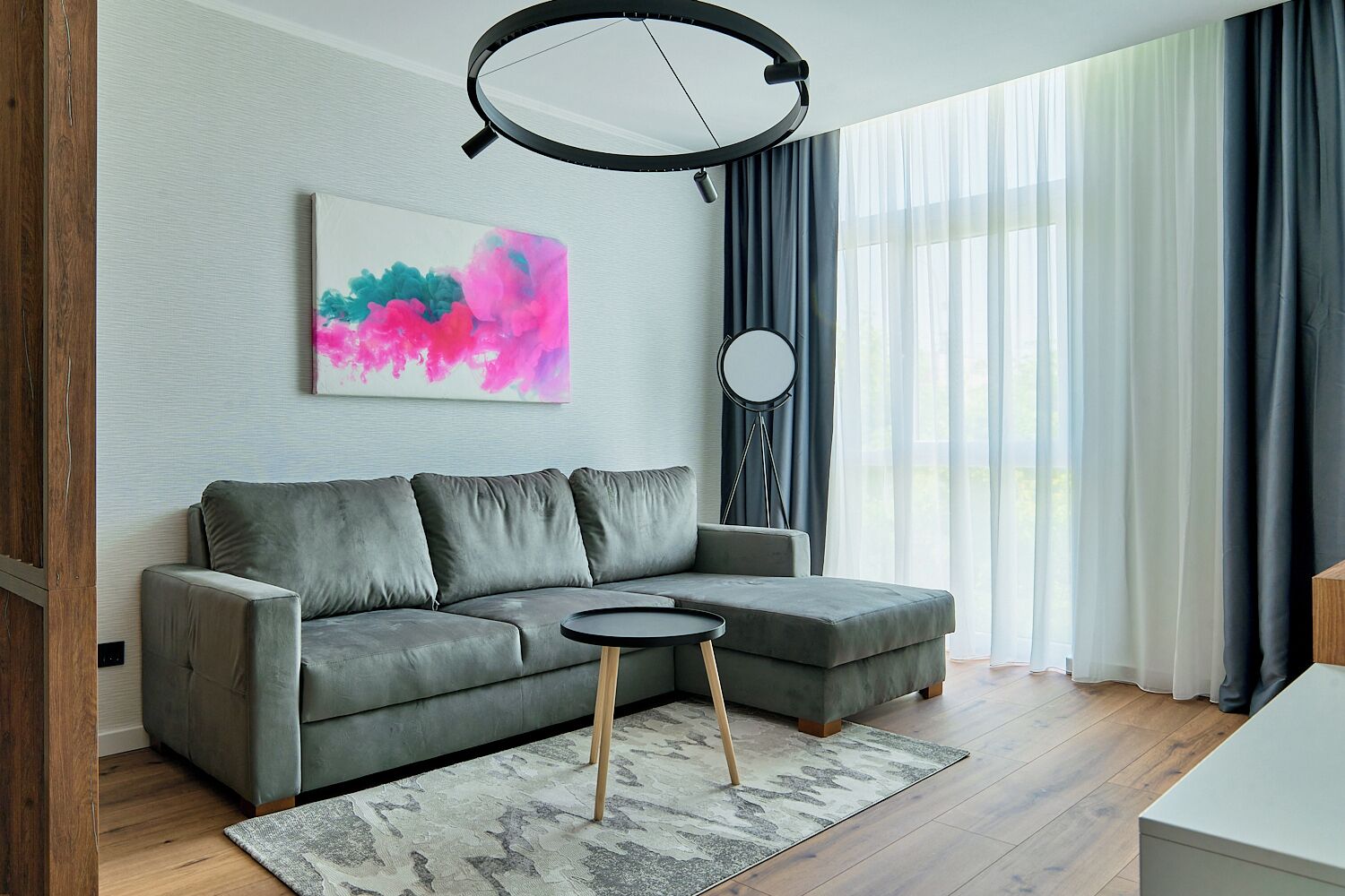 Un salon moderne avec un canapé d’angle gris, une petite table ronde et un grand tableau abstrait accroché au mur.