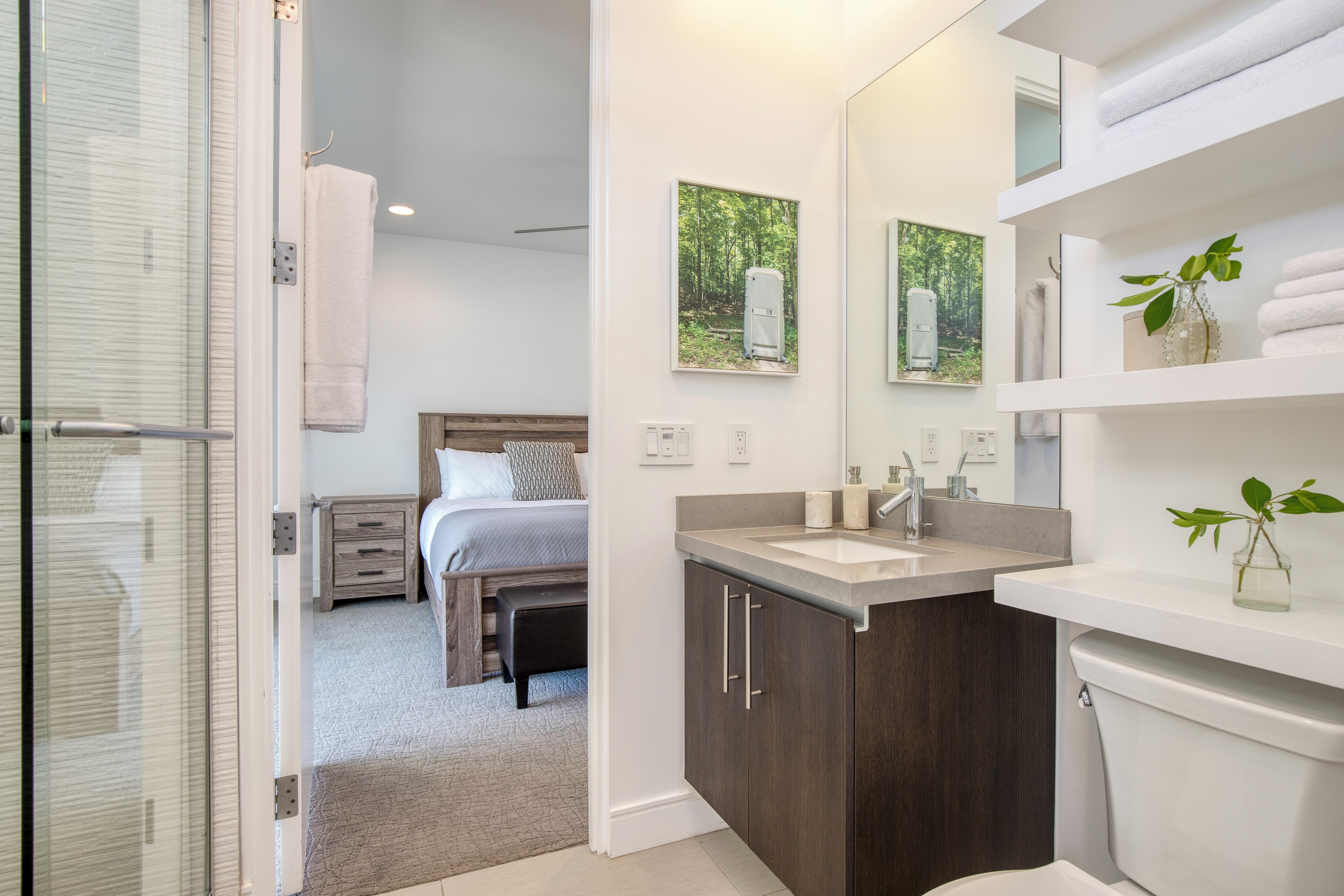 A modern bathroom with a large mirror, a sink, and a toilet. There is a bed in the adjacent room.