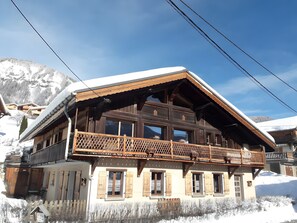 A wooden chalet with a balcony, large windows, and a snowy mountain backdrop.