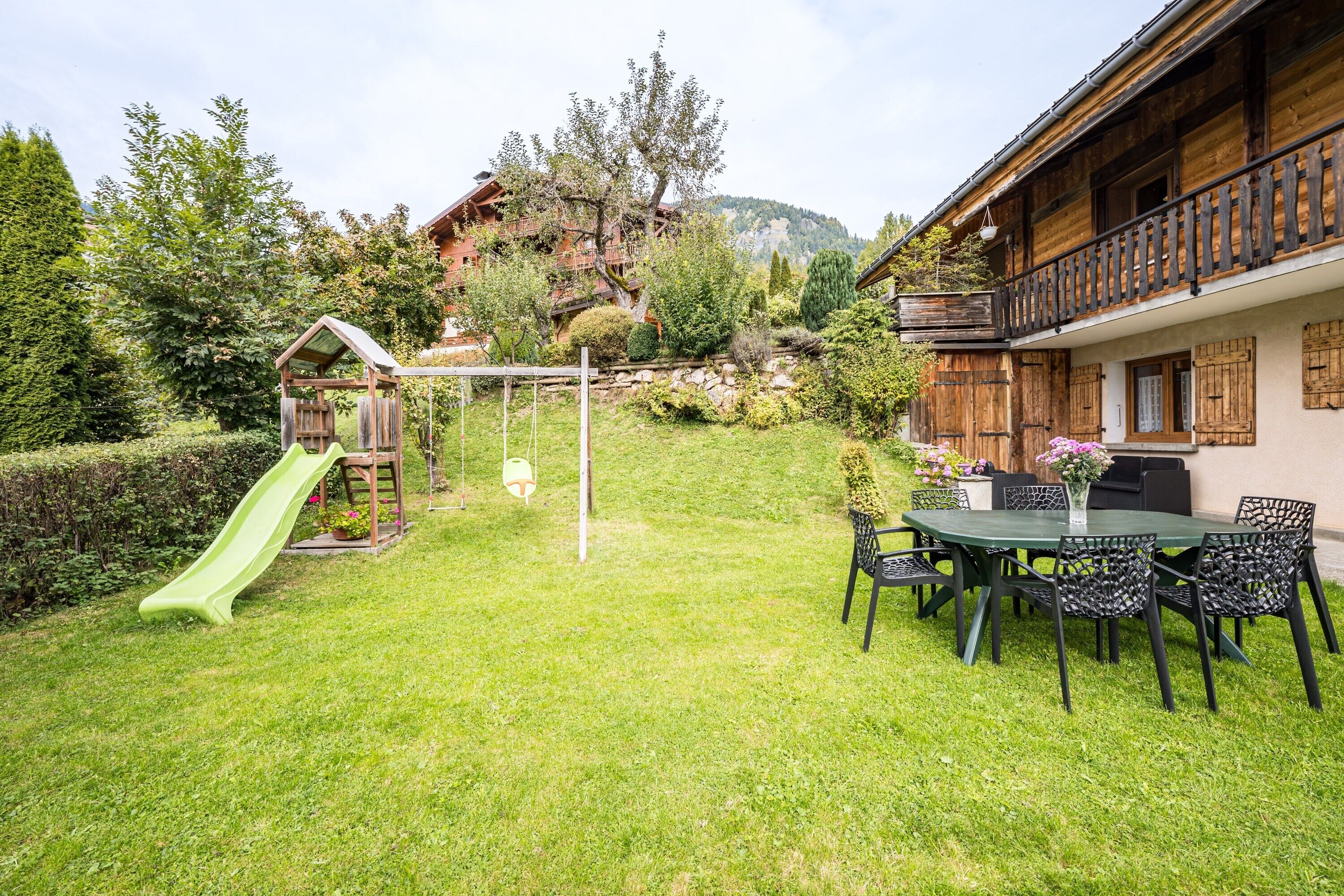 A wooden house with a balcony, a green slide, and a dining area with chairs and a table.