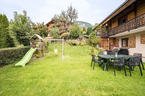 Una casa de madera con balcón, un tobogán verde y una zona de comedor con sillas y mesa.