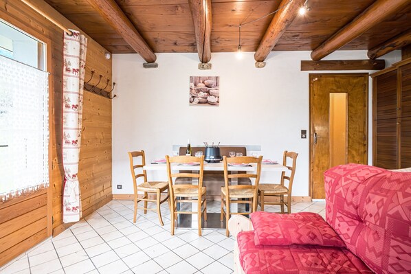 A dining area with a wooden table and chairs, a kitchen in the background, and a red patterned sofa.