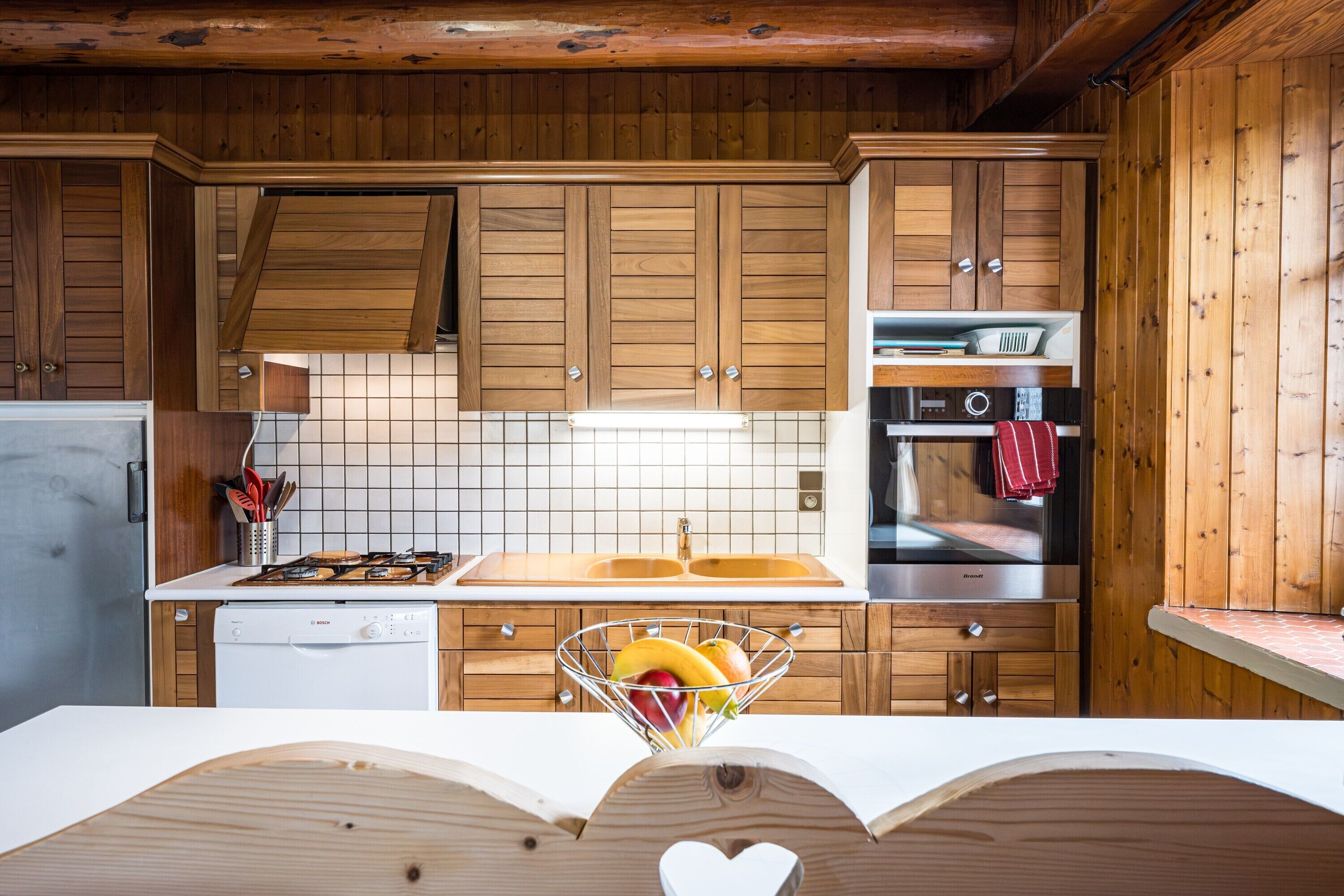 Une cuisine avec des meubles de rangement en bois, un plan de travail blanc, un évier en acier inoxydable et un lave-vaisselle.