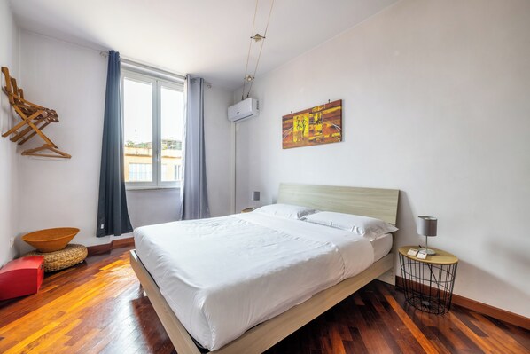 A modern bedroom with a wooden bed, a round side table, a wall-mounted air conditioner, and a large window with curtains.