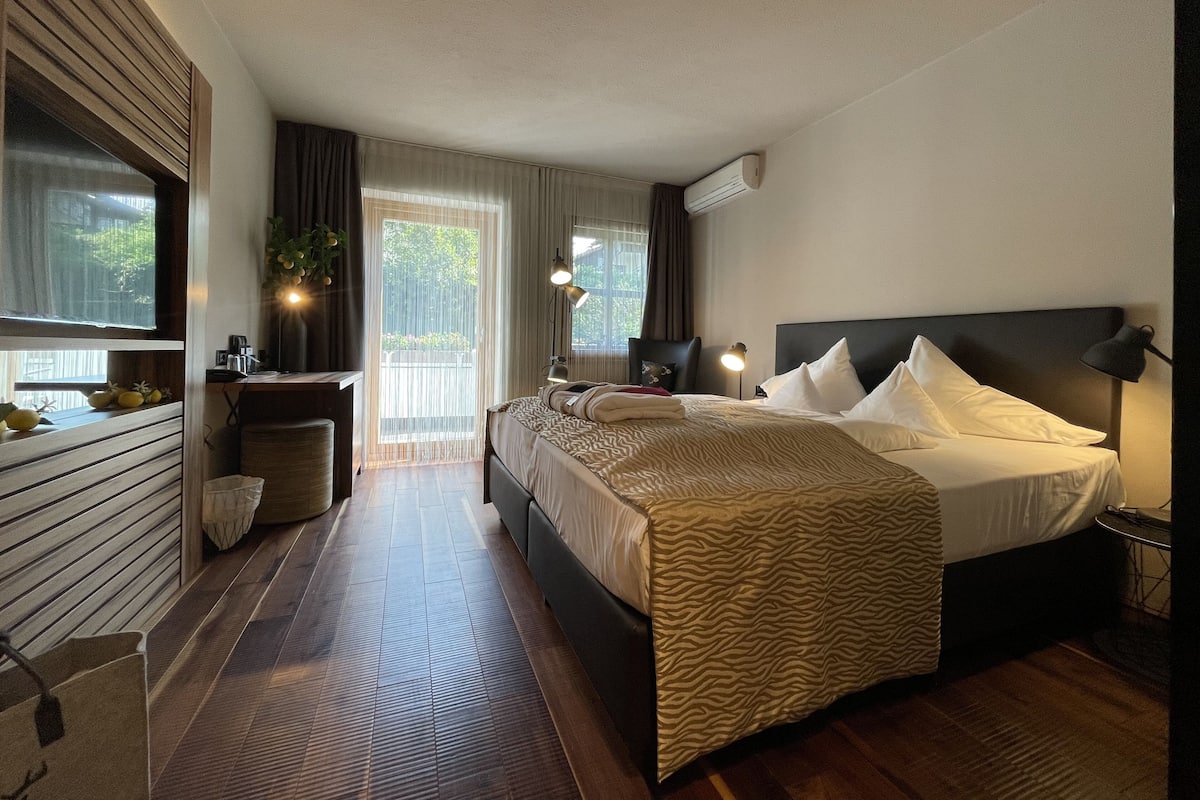 A modern hotel room with a large bed, a desk with a chair, a small table, and a window with curtains.
