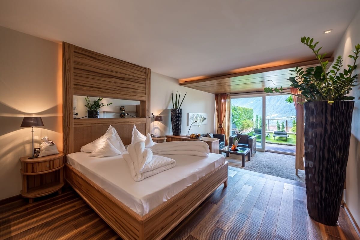 A modern hotel room with a large bed, wooden headboard, a sitting area with a sofa and coffee table, and a large window offering a view of the outdoors.
