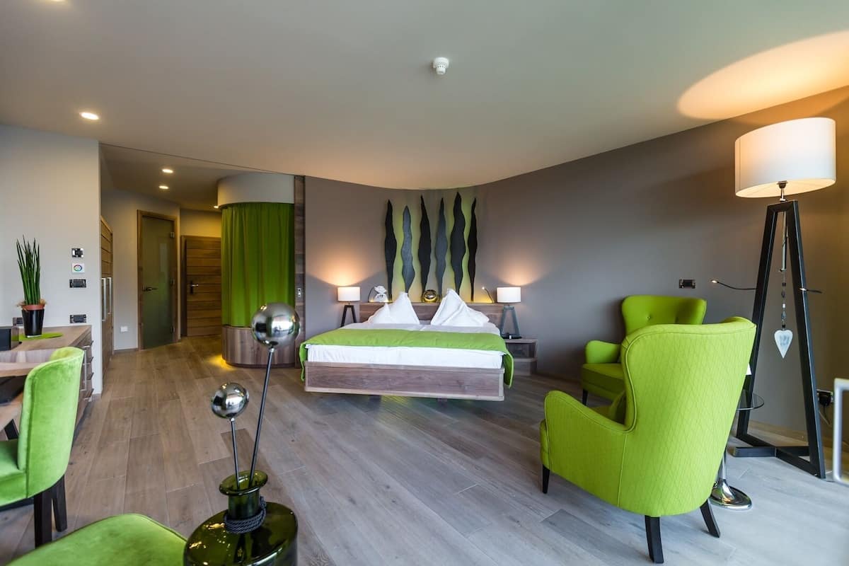 A modern hotel room with a bed, green chairs, a desk, and a lamp.