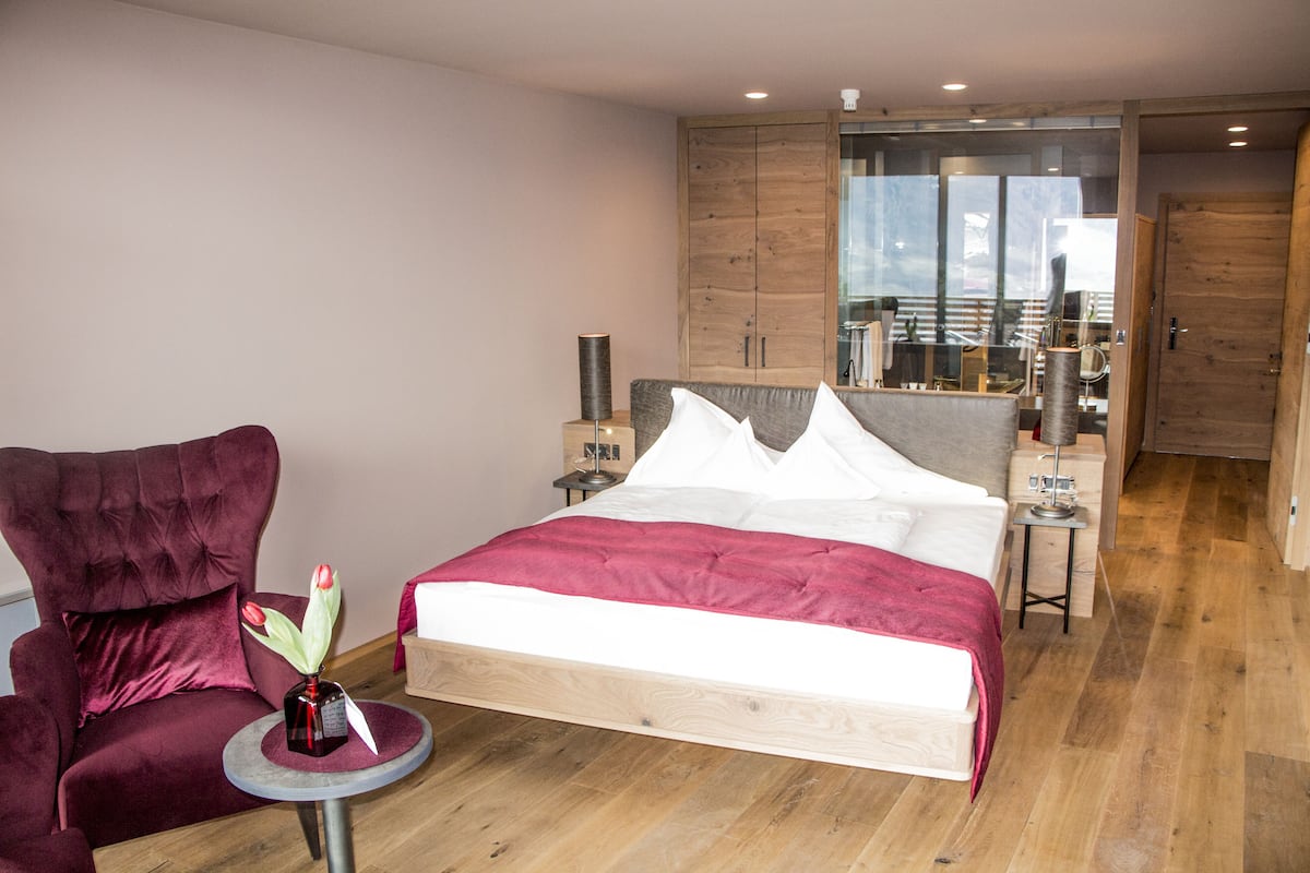 A modern hotel room with a large bed, a red armchair, a small round table with a vase, and wooden flooring.