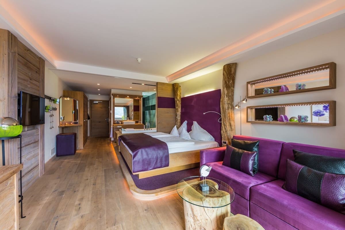 A modern hotel room with a large bed, a purple sofa, a glass coffee table, and a wooden floor.