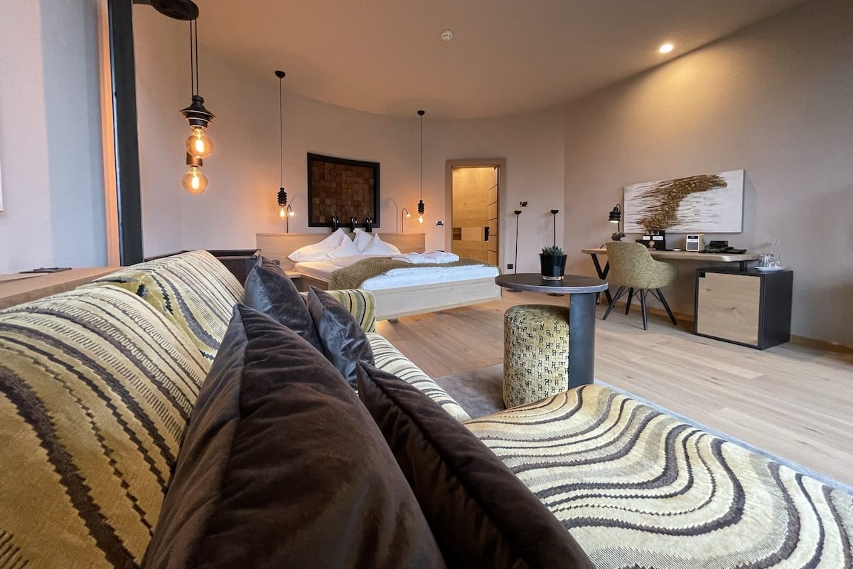 A modern hotel room with a large bed, a desk, and a chair. There are multiple light fixtures and a painting on the wall.