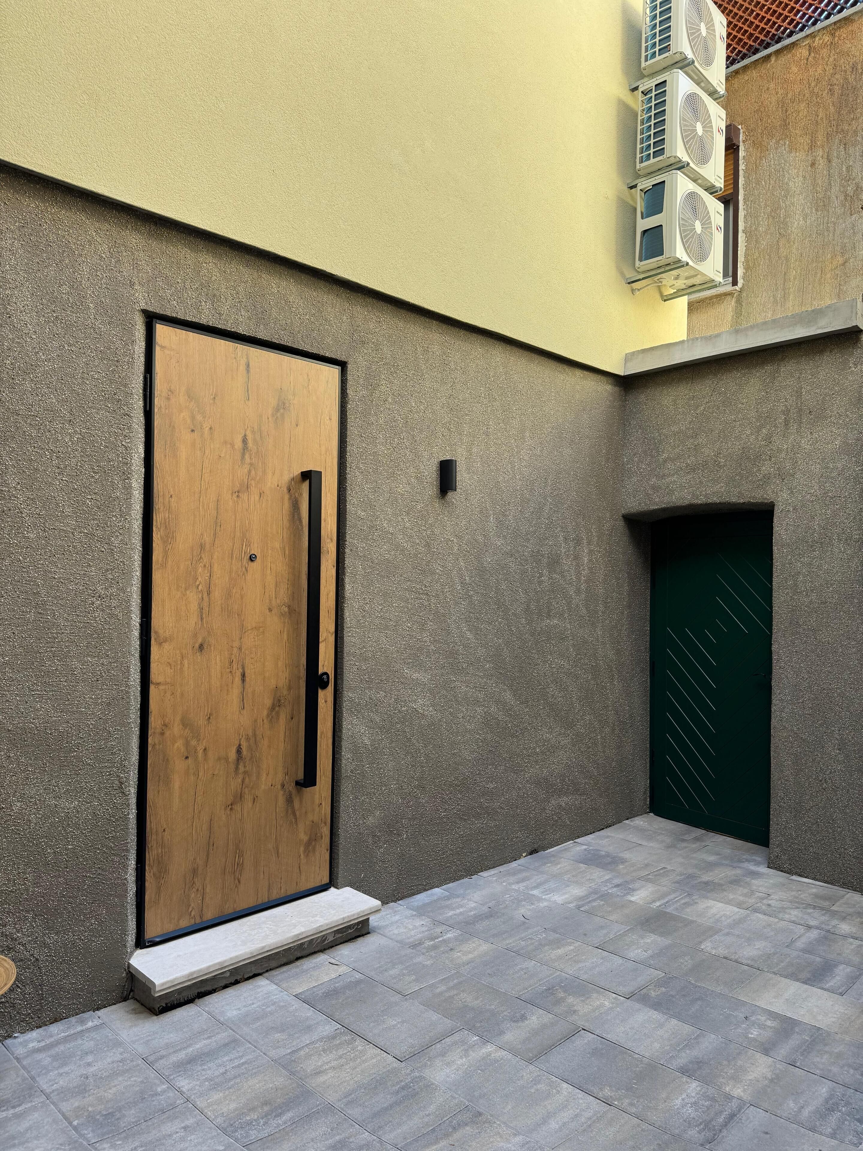 Ein Innenhof mit einer Holztür und einer grünen Tür, eine kleine Stufe führt zur Tür, und Klimaanlagen sind an der Wand angebracht.