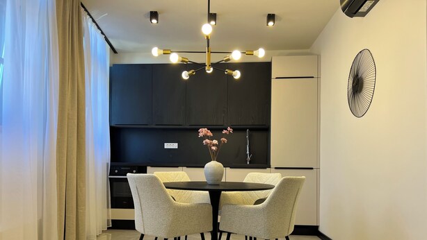 A modern dining area with a round table, two chairs, a vase with flowers, a wall-mounted fan, and a kitchen in the background.