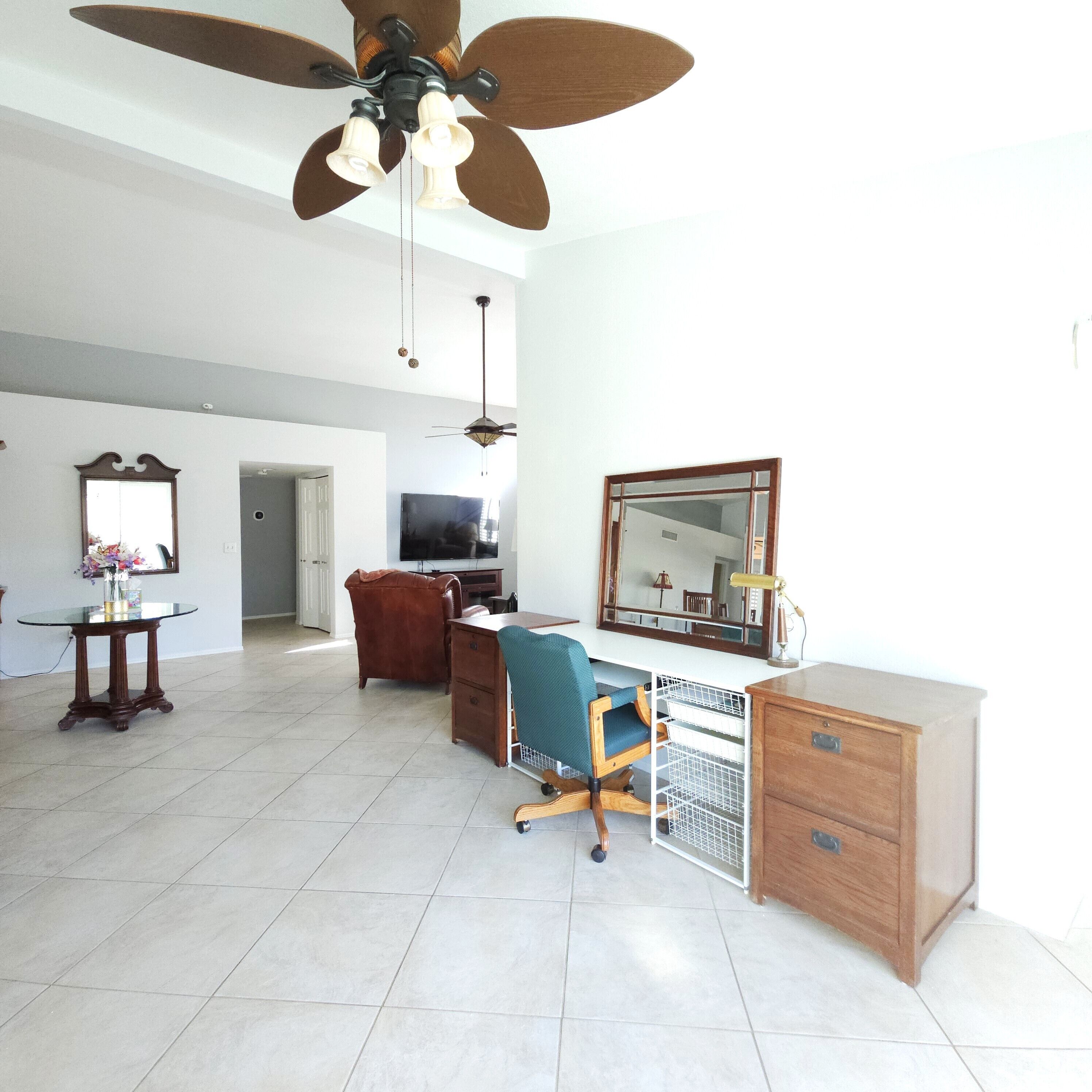 A spacious living room with a ceiling fan, a television, a desk with a chair, and a large mirror.